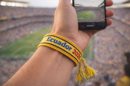 Hand wearing a bracelet with 'Ecuador 2020' text, taking a photo of a stadium.